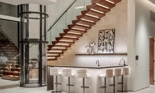 Round glass elevator installed alongside a brightly lit staircase and bar area.