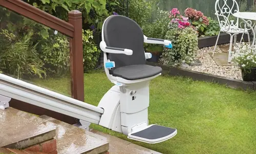 A Handicare 1000 stairlift stationed at the bottom landing in a backyard garden setting.