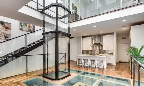 An octagonal glass elevator installed along a black staircase on top of transparent glass floor.
