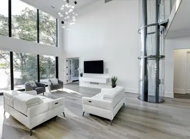 Modern luxury living room with panoramic glass residential elevator, open‑concept design, floor‑to‑ceiling windows, white furniture, and hardwood flooring.