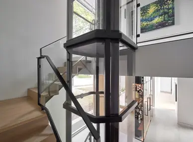 Panoramic glass residential elevator with black square frame installed at a stair landing, featuring transparent walls, modern railing, and open‑concept luxury home interior.