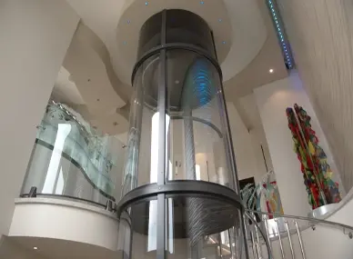 Panoramic glass residential elevator with cylindrical design installed in a luxury home atrium, featuring curved balconies and modern architectural interior.