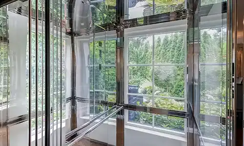 Glass panoramic home elevator with reflective metal framing, shown next to large windows overlooking dense green trees.