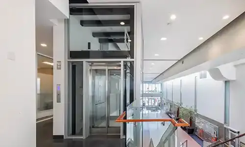 Glass elevator with metal framing located on an upper floor of a bright commercial building, beside a glass railing overlooking the level below.