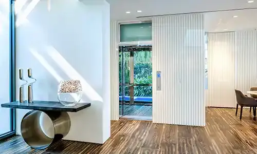 Home elevator with glass doors in a bright modern interior, surrounded by wood‑grain flooring and white paneled walls.
