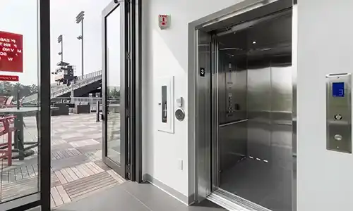 Stainless‑steel elevator with open doors inside a stadium facility, positioned next to large glass doors leading out to the seating area.
