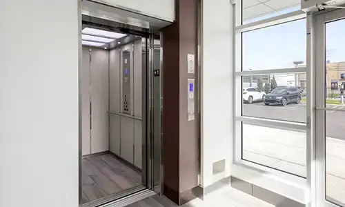 Commercial elevator with open stainless steel doors, modern interior, illuminated control panel, and large windows in a professional office building lobby.