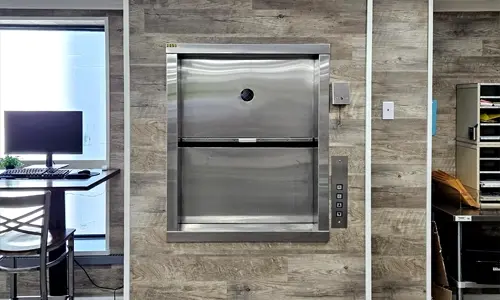 Material lift with a stainless‑steel front door set into a wood‑textured wall, featuring a vertical sliding door panel, call buttons on the right side, and a compact workstation visible to the left.