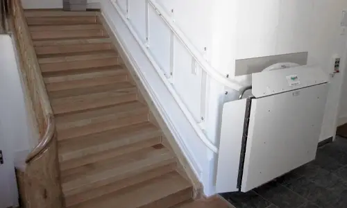 An inclined curved platform lift folded while not in use at the bottom landing of staircase.