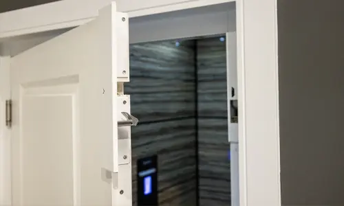 Door interlock bracket mounted on the inside edge of a swing door, shown with an open doorway leading into a residential elevator cab with wood‑textured interior panels.
