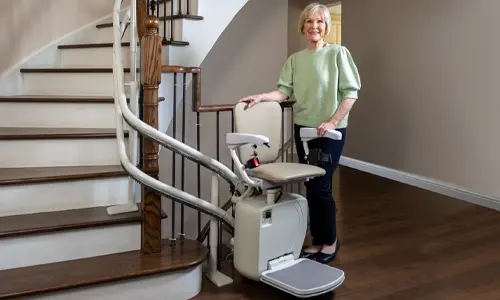 Stairlift positioned on a curved staircase with a person standing beside the chair, showing the rail, seat, and footrest in a home interior.