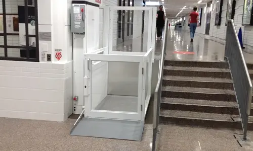 White-framed, unenclosed vertical platform lift installed next to a flight of stairs in an institutional setting.