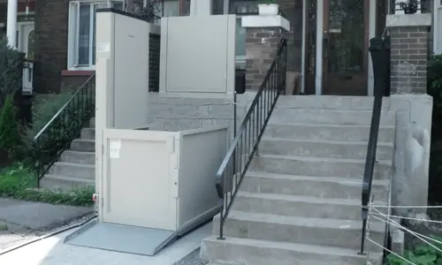 Outdoor vertical platform lift installed beside a front staircase at a residential entrance.