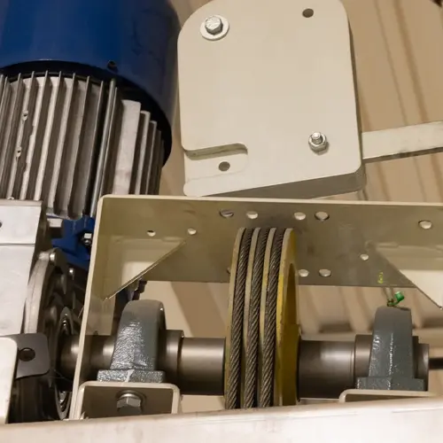 Close-up of an elevator drive assembly showing a grooved sheave with multiple hoist cables wrapped around it, supported by two bearing housings on a horizontal shaft, with a motor and mounting brackets visible above the components.