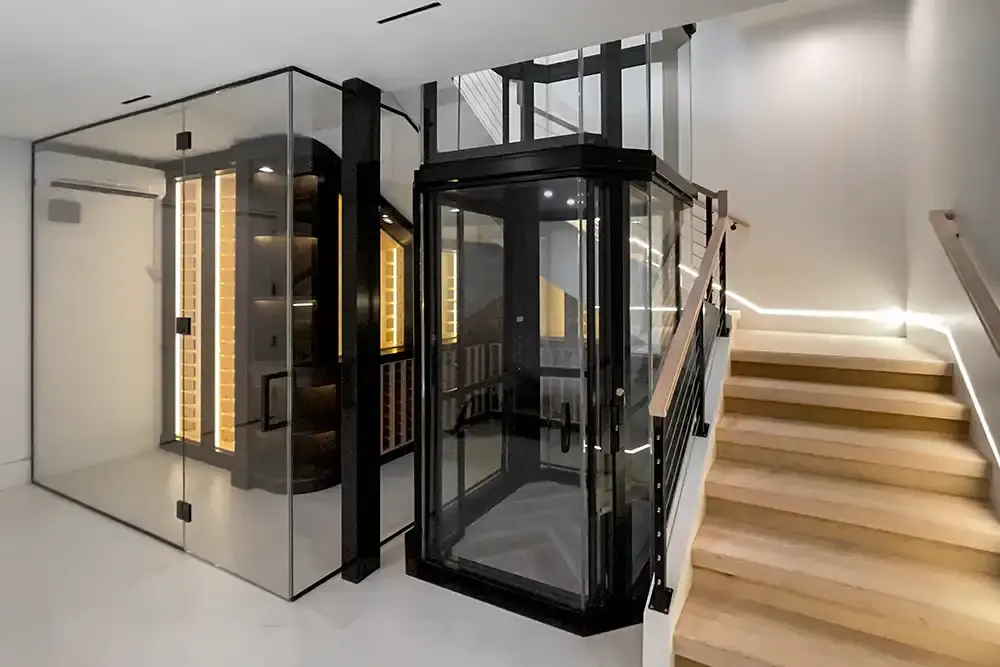 Glass residential elevator with a black frame positioned beside a modern staircase, shown next to a glass‑enclosed wine room with illuminated shelving.