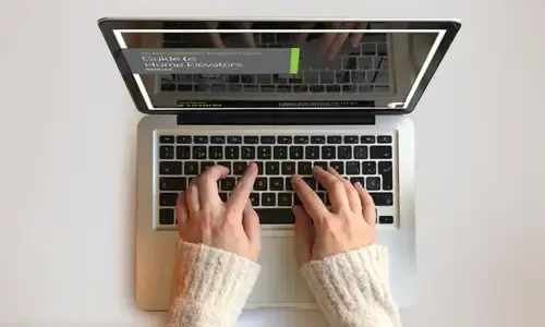 Hands typing on a laptop keyboard, with the screen displaying a webpage titled “Guide to Home Elevators.”