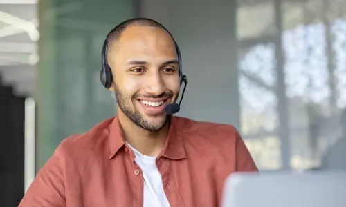 Person wearing a headset and a rust‑colored shirt seated at a desk, working on a laptop in a modern office environment.