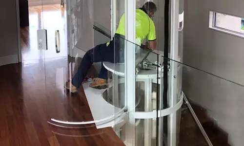 Elevator technician servicing a panoramic glass residential elevator, performing maintenance inside a cylindrical home lift installed in a modern multi‑level interior.