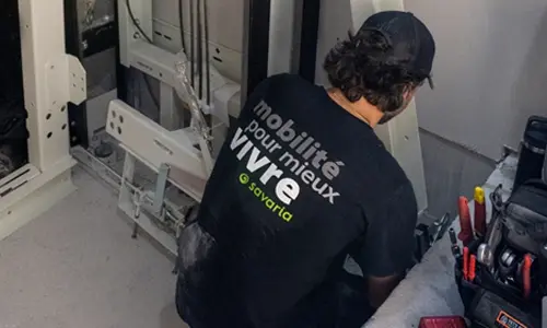 Elevator technician installing and servicing residential home elevator components inside a lift shaft, wearing Savaria mobility workwear and using professional tools.