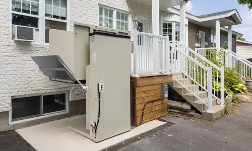 Outdoor vertical platform lift with weatherproof enclosure installed beside a residential porch, providing wheelchair‑accessible entry from driveway to main floor.