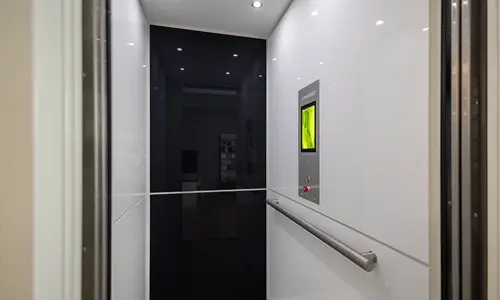 Modern residential elevator interior with glossy white and black wall panels, stainless steel handrail, illuminated touchscreen control panel, and recessed LED ceiling lights.