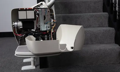 Residential stairlift with seat cover removed, exposing internal motor, wiring, and mechanical components during maintenance on a carpeted staircase.