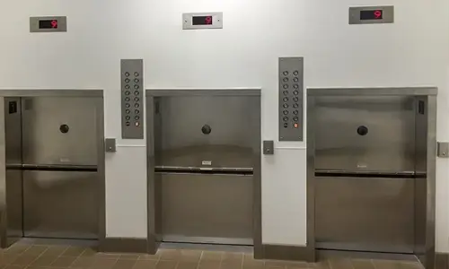 Stainless steel material lifts with vertical sliding doors and wall‑mounted call button panels installed in a commercial building for transporting goods between floors.