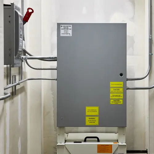 Savaria elevator controller cabinet mounted in a mechanical room, featuring gray metal enclosure, warning labels, and electrical conduit connections.