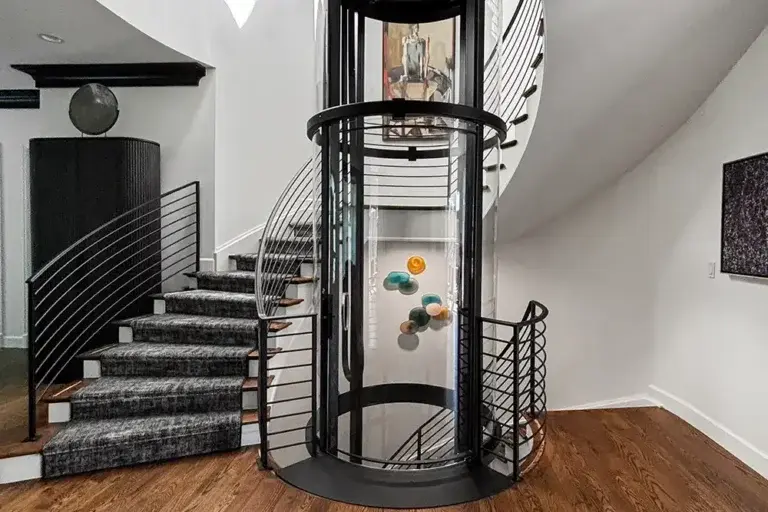 Panoramic glass residential elevator with curved black frame installed between sweeping staircases in a modern luxury home with hardwood floors.