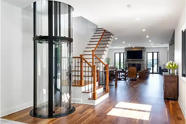 Panoramic glass residential elevator with cylindrical design installed in a modern open‑concept home, featuring hardwood floors, staircase, and bright living space.