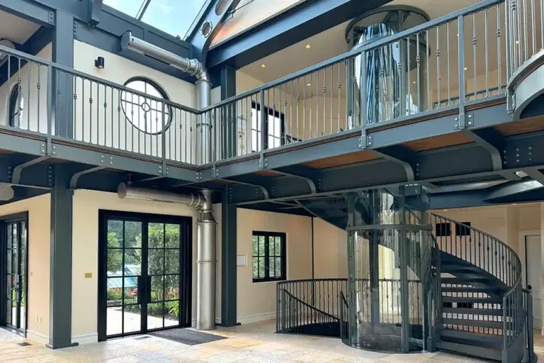 Industrial‑style luxury home interior featuring a full‑height panoramic glass elevator, exposed steel beams, spiral staircase, and open mezzanine levels.
