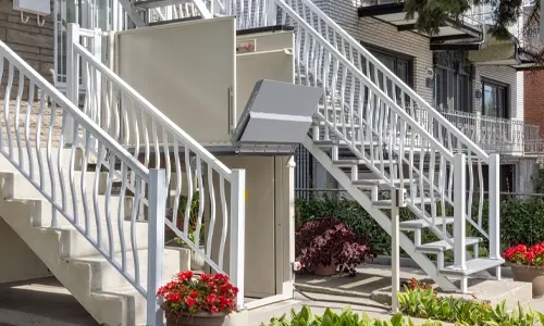 Outdoor vertical platform lift with beige metal enclosure installed between exterior staircases of a residential building, providing wheelchair and mobility accessibility from ground level to an elevated entrance.