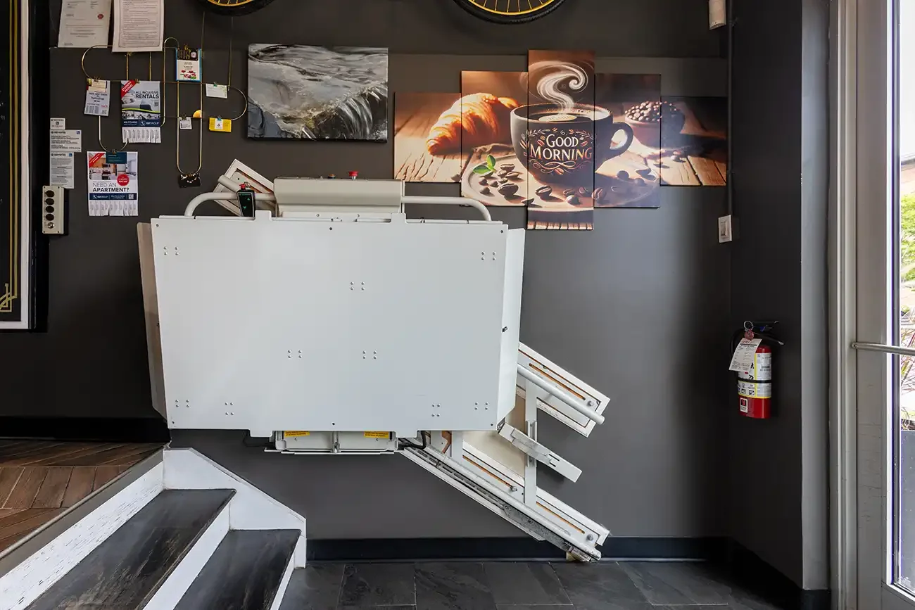 Savaria Delta inclined platform lift parked at a commercial stair landing, featuring a folded wheelchair platform, wall‑mounted rail system, and compact design to maintain clear stairway access.