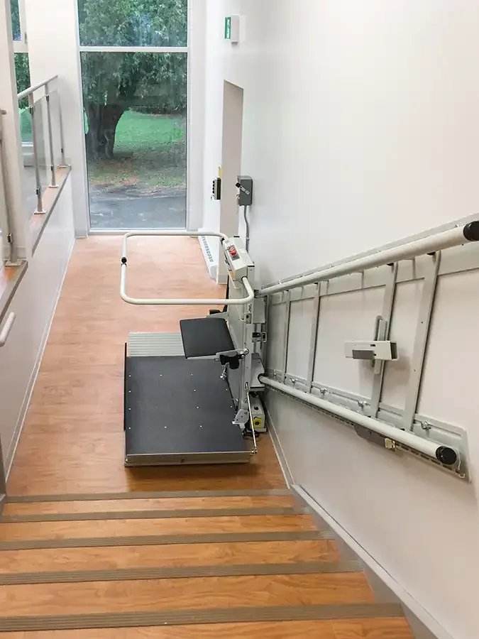 Savaria Delta inclined platform lift at upper landing, showing fold‑down wheelchair platform, safety arms, and wall‑mounted rail system installed indoors for commercial accessibility.