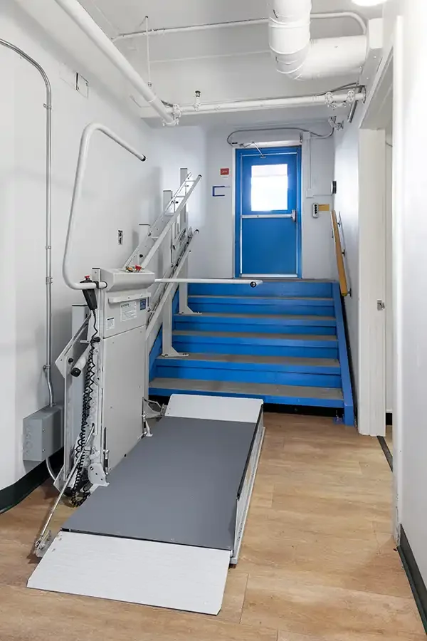 Savaria Delta inclined platform lift deployed at the base of a commercial staircase, featuring a full‑size wheelchair platform, safety arms, and wall‑mounted rail system for accessible travel between floor levels.