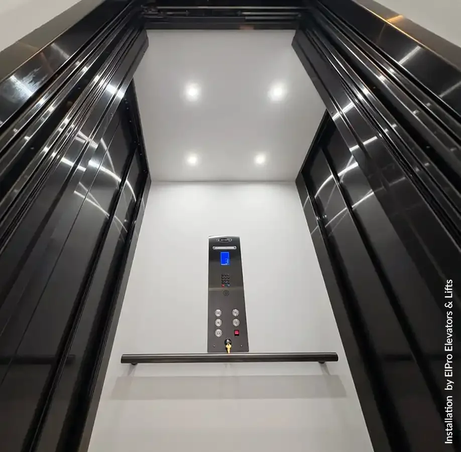 Residential elevator interior looking upward with glossy white ceiling, recessed LED lights, stainless steel side panels, modern control panel, and horizontal handrail inside a luxury home elevator cabin.