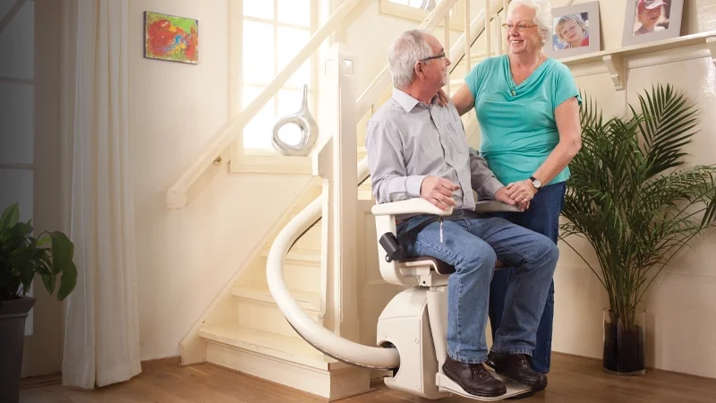 A smiling couple riding a single-rail curved stairlift inside their home.