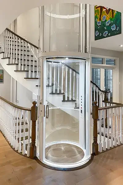 Round glass residential elevator with a white frame positioned at the base of a curved staircase in a bright, modern home interior.