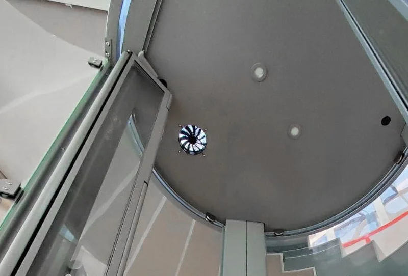 Ceiling view from inside a glass home elevator, showing the round cab roof with two recessed lights and a small ventilation fan at the center, framed by the curved glass walls and open door below.