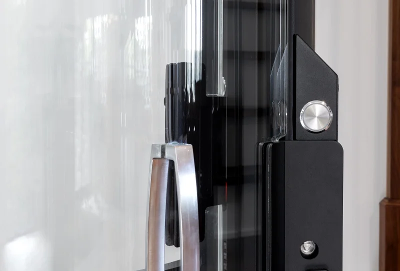 Close‑up of a glass elevator door handle and latch mechanism, featuring a metal pull handle, a black frame, and a round stainless‑steel release button beside the transparent door panel.