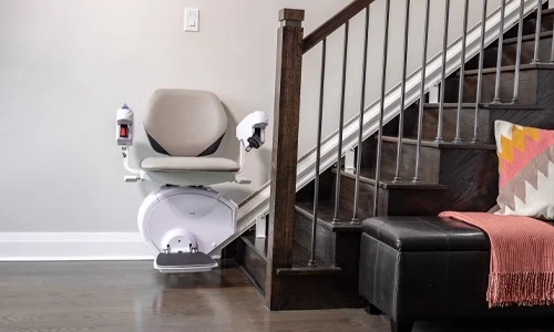 Stairlift parked at the bottom of a wooden staircase, featuring a padded seat, armrests, and footrest mounted on a rail along the side of the stairs in a modern home interior.
