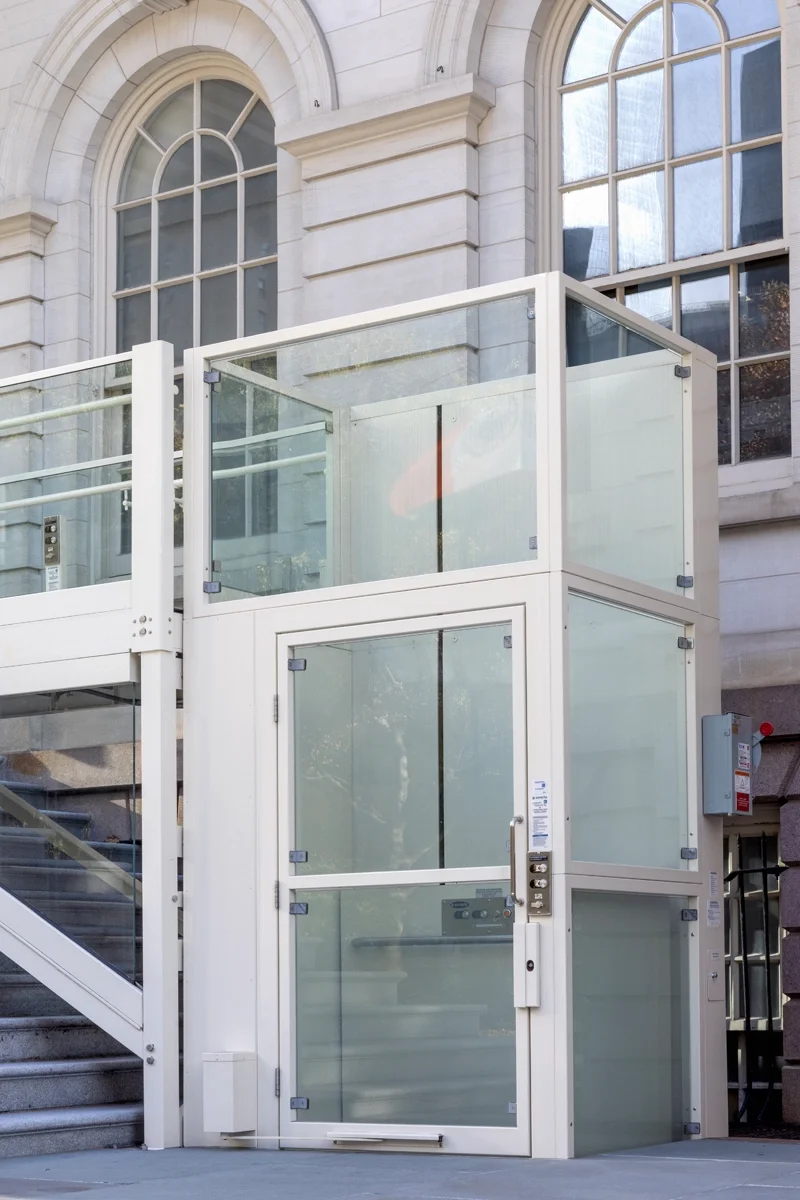 Outdoor vertical platform lift with a white frame and frosted glass panels, installed beside a stone staircase and positioned against a building with large arched windows.