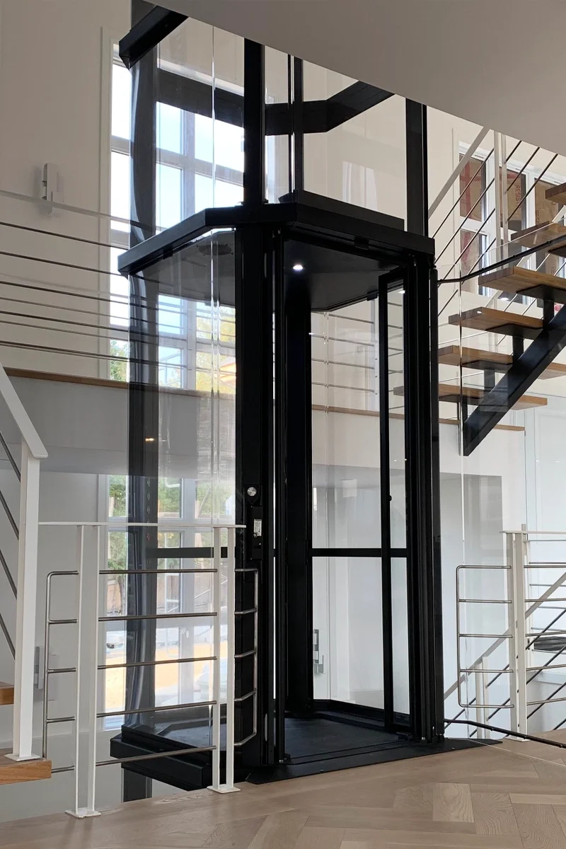 Glass‑enclosed home elevator with a black frame, positioned between staircases and surrounded by modern railings in a bright, open interior space.