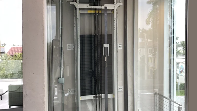 Elevator hoistway with exposed guide rails, chains, and mechanical components enclosed in glass, showing the lift structure within a modern home.