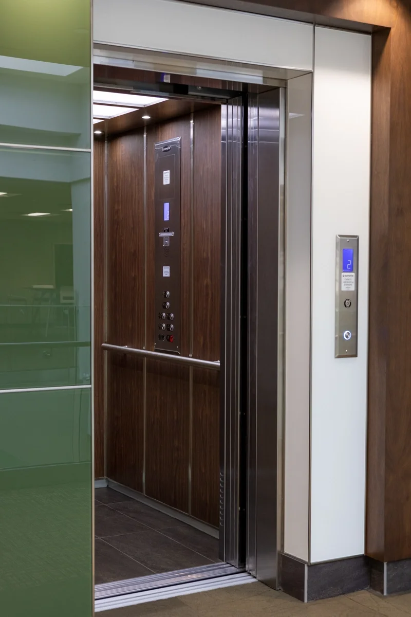 Modern commercial elevator with open stainless steel doors, wood‑paneled interior, illuminated control panel, handrail, and wall‑mounted call button in a professional building interior.
