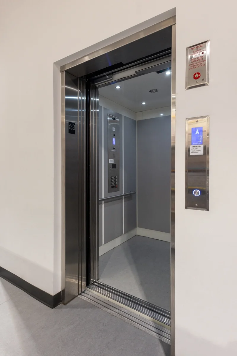 Commercial elevator with stainless steel door frame, illuminated interior control panel, and wall-mounted call buttons in a modern office building hallway.