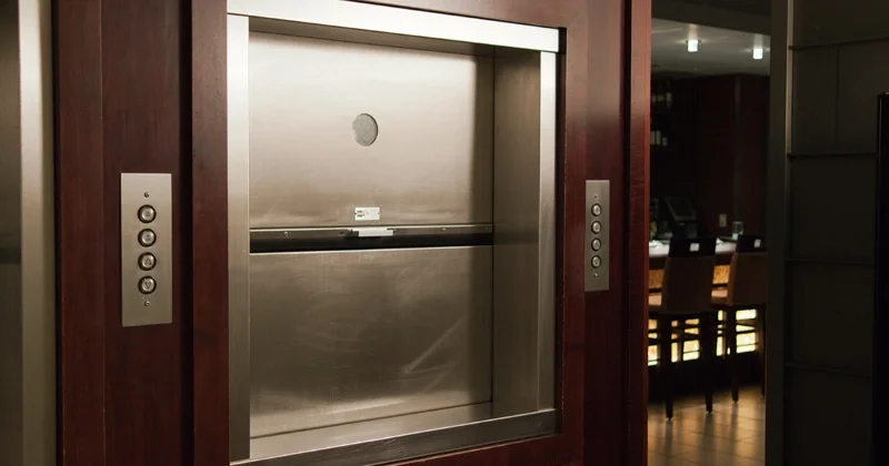 Stainless steel dumbwaiter with closed vertical sliding door and wall‑mounted call buttons, installed in a commercial or residential interior for transporting food and items between floors.