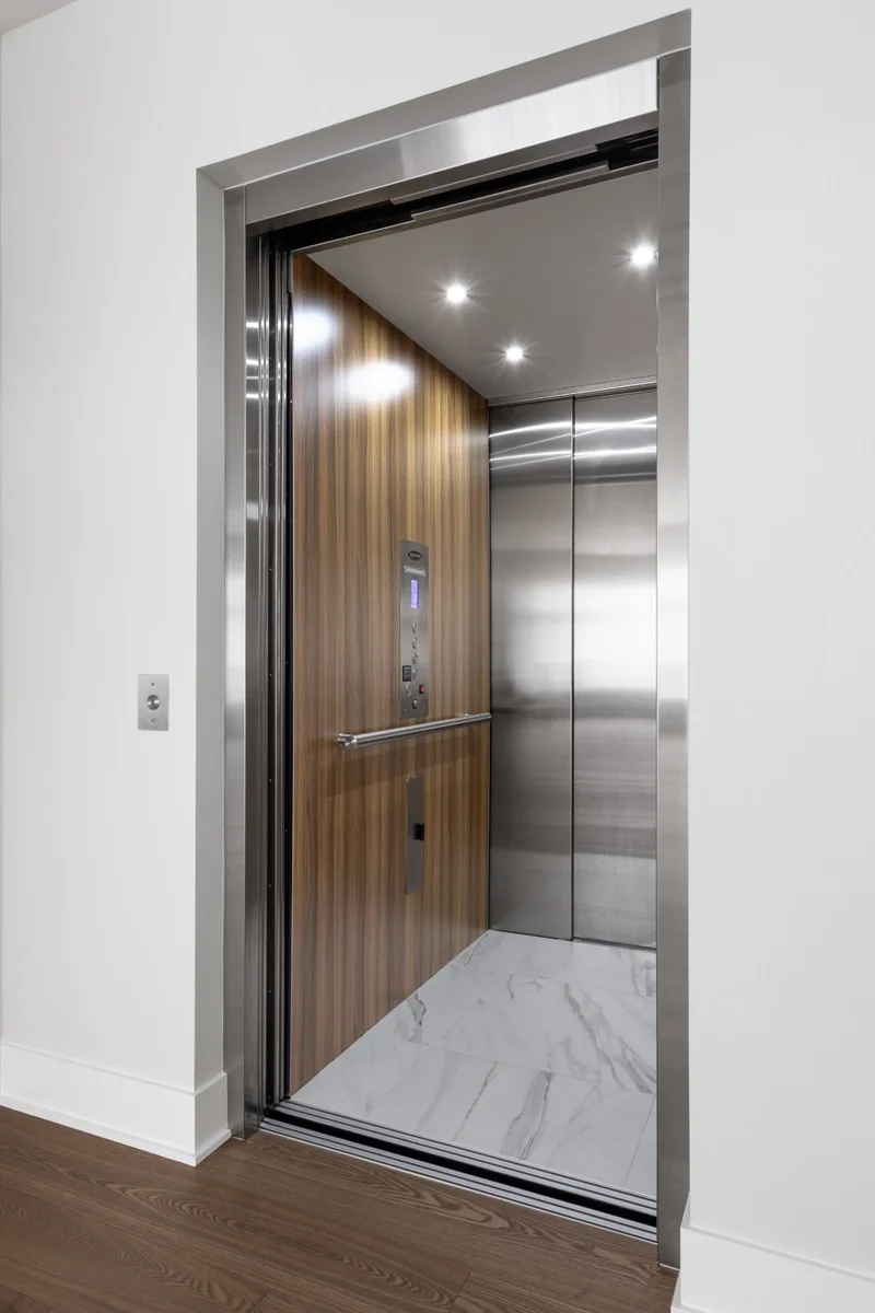 Luxury home elevator with stainless steel trim, wood‑grain interior walls, illuminated control panel, handrail, and marble‑look flooring installed in a modern residential home.