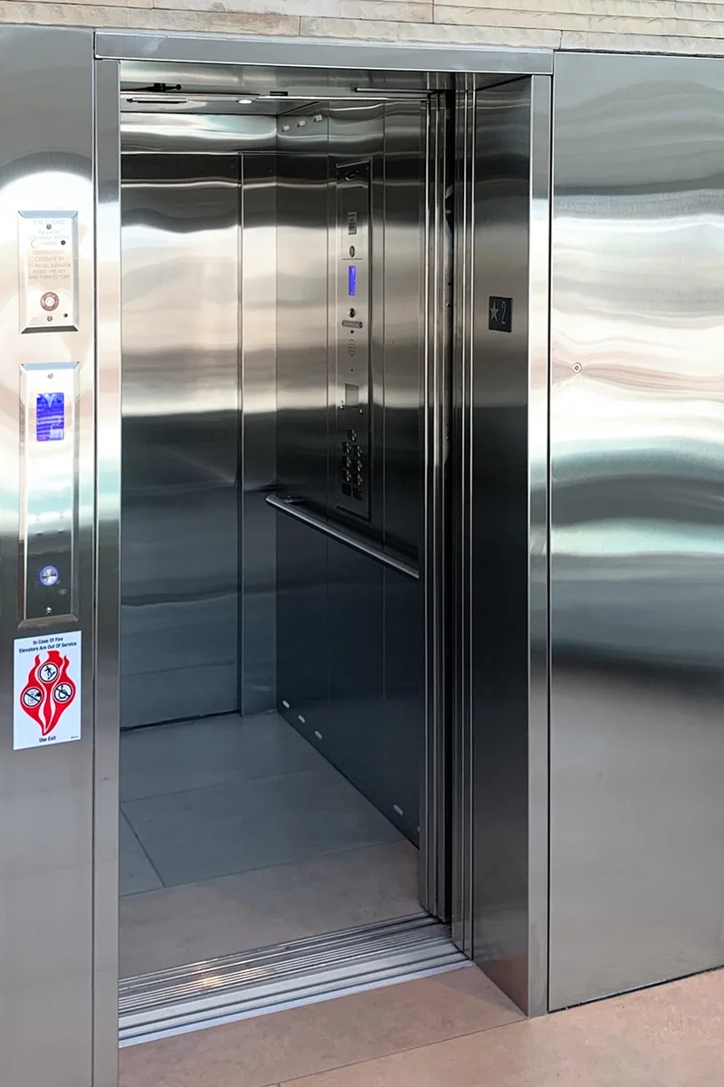 Commercial stainless steel elevator with open doors, illuminated control panel, call stations, and modern interior installed in a public or commercial building.