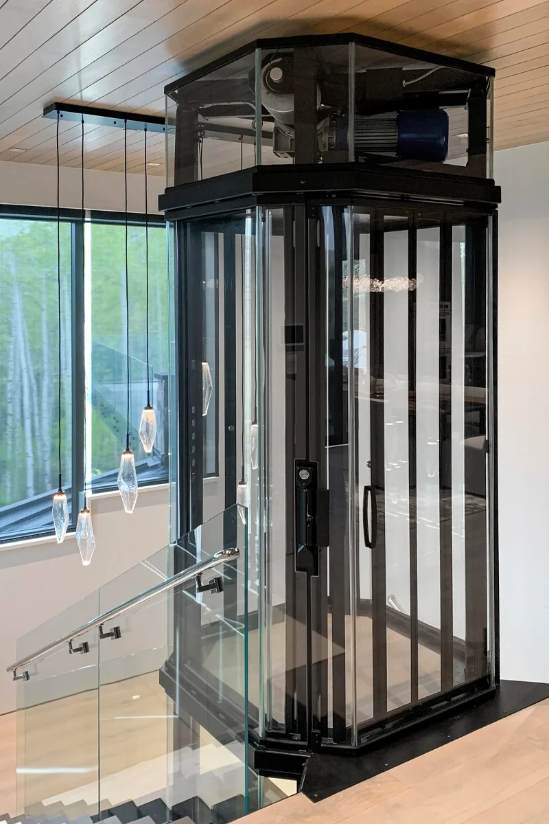 Panoramic glass residential elevator with black steel frame and transparent cabin, installed beside a staircase in a modern luxury home.
